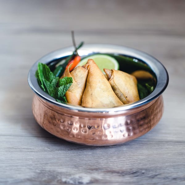 Golden-brown samosas garnished with mint, lime, and chili in a hammered copper bowl.