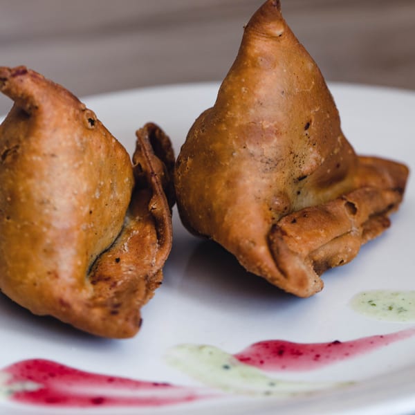 Two golden-brown samosas on a white plate, garnished with red and green sauces.