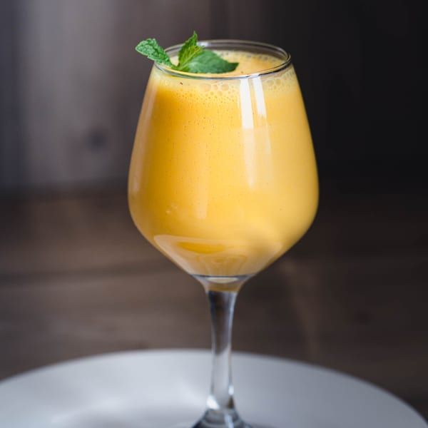 A vibrant yellow mango lassi in a wine glass, garnished with fresh mint leaves, on a white plate.